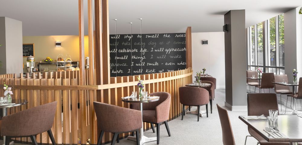 Modern cafe interior with brown chairs and round tables on a light carpet. A blackboard displays inspirational quotes in white script, adding a cozy vibe.