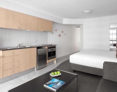 Minimalist studio apartment with light wood kitchenette, stainless steel appliances, and a bed. A sleek coffee table holds green apples and a blue book.