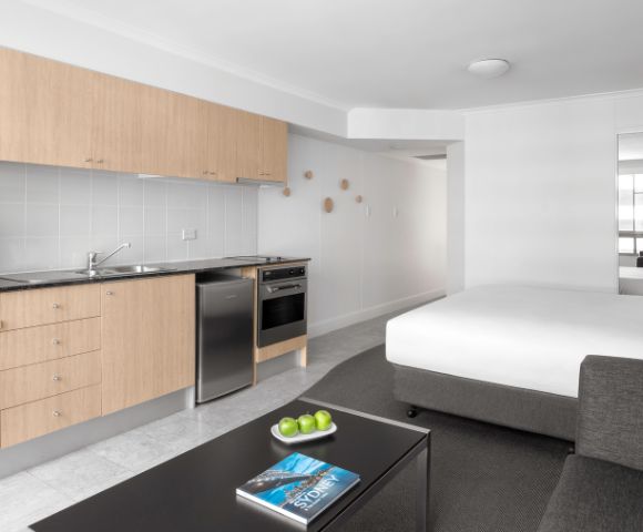 Minimalist studio apartment with light wood kitchenette, stainless steel appliances, and a bed. A sleek coffee table holds green apples and a blue book.