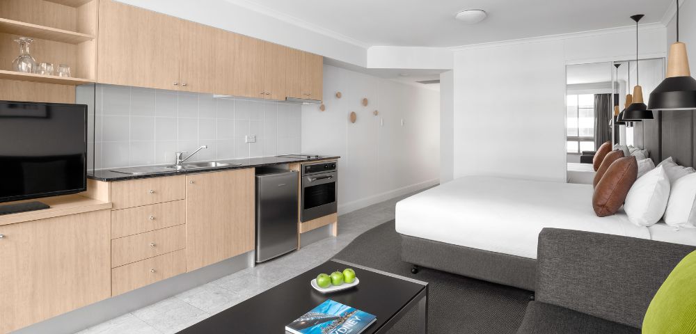 Minimalist studio apartment with light wood kitchenette, stainless steel appliances, and a bed. A sleek coffee table holds green apples and a blue book.