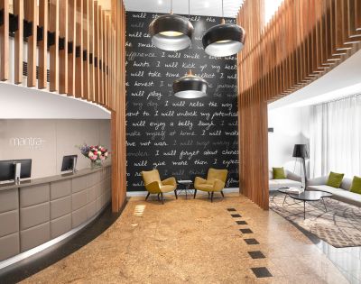 Modern hotel lobby with a curved reception desk on the left, a cozy seating area with two yellow chairs, a dark feature wall with inspirational text, and hanging lights.
