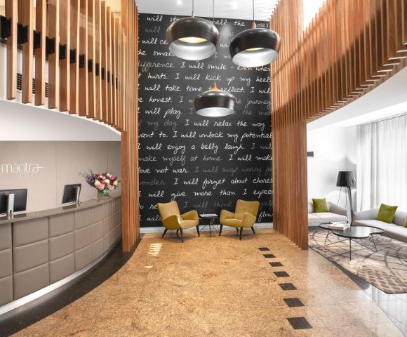 Modern hotel lobby with a curved reception desk on the left, a cozy seating area with two yellow chairs, a dark feature wall with inspirational text, and hanging lights.