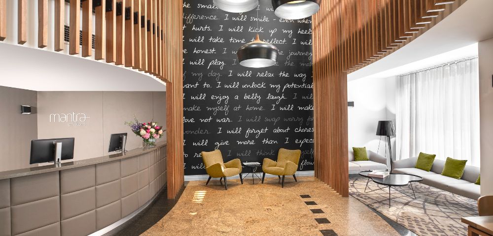 Modern hotel lobby with a curved reception desk on the left, a cozy seating area with two yellow chairs, a dark feature wall with inspirational text, and hanging lights.
