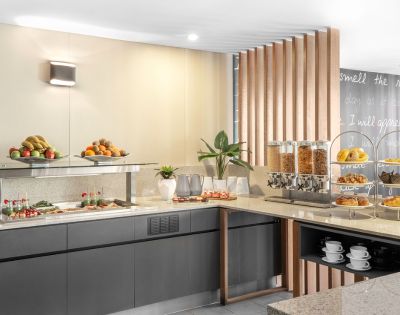 Contemporary buffet with fresh fruits, pastries, cereal dispensers, and neatly stacked cups. Warm lighting and sleek decor create an inviting atmosphere.