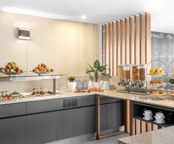 Contemporary buffet with fresh fruits, pastries, cereal dispensers, and neatly stacked cups. Warm lighting and sleek decor create an inviting atmosphere.