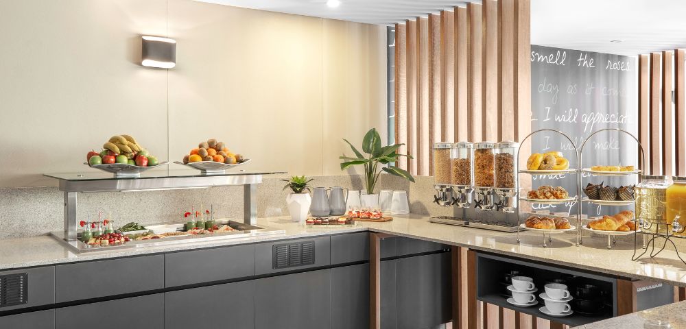Contemporary buffet with fresh fruits, pastries, cereal dispensers, and neatly stacked cups. Warm lighting and sleek decor create an inviting atmosphere.