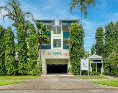 Modern three-story building with green balconies, surrounded by tall palm trees under a clear blue sky. Entrance leads to a shaded driveway.