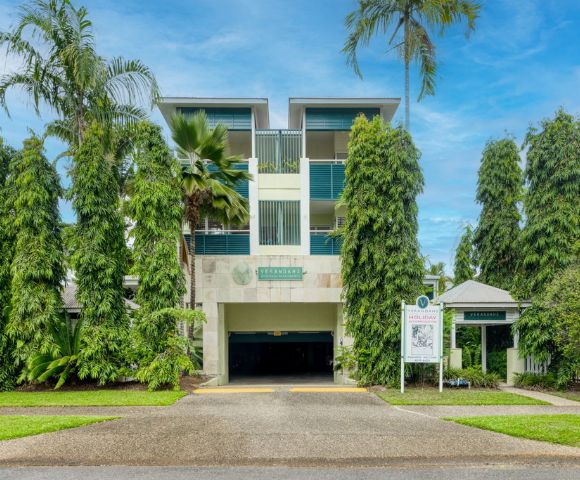 Modern three-story building with green balconies, surrounded by tall palm trees under a clear blue sky. Entrance leads to a shaded driveway.