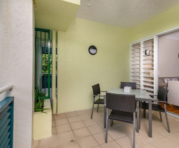 A cozy patio with a glass table surrounded by four wicker chairs, next to a sliding door leading inside. The soft yellow walls and potted plant add a welcoming feel.