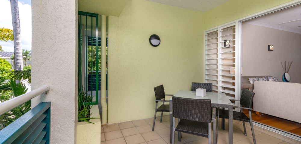 A cozy patio with a glass table surrounded by four wicker chairs, next to a sliding door leading inside. The soft yellow walls and potted plant add a welcoming feel.