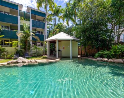 Clear turquoise pool with a small gazebo, surrounded by palm trees and garden, adjacent to a modern apartment building, conveying a relaxed, tropical atmosphere.