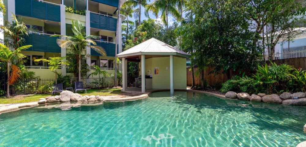 Clear turquoise pool with a small gazebo, surrounded by palm trees and garden, adjacent to a modern apartment building, conveying a relaxed, tropical atmosphere.