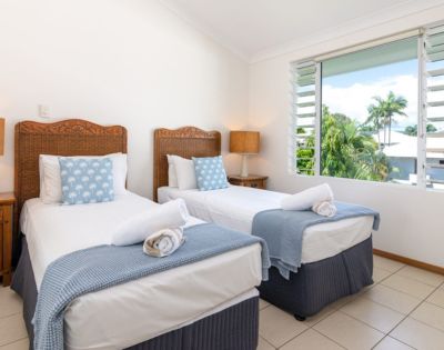 Two neatly made twin beds with wicker headboards, blue bedding, and polka-dot cushions in a bright room with tile floors and a window view of trees.