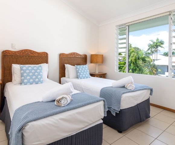 Two neatly made twin beds with wicker headboards, blue bedding, and polka-dot cushions in a bright room with tile floors and a window view of trees.
