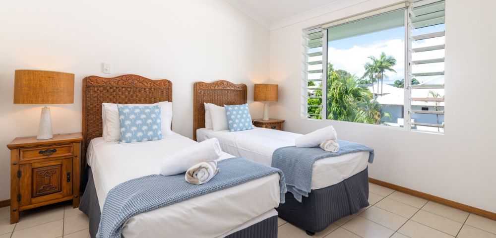 Two neatly made twin beds with wicker headboards, blue bedding, and polka-dot cushions in a bright room with tile floors and a window view of trees.