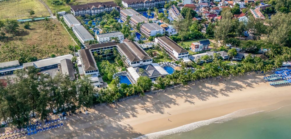 Aerial view of a coastal resort with white sand beach and turquoise sea, surrounded by lush greenery and buildings, evoking a serene and inviting atmosphere.