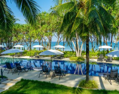 Tropical poolside oasis with palm trees and white umbrellas, overlooking a serene blue ocean. Relaxing lounge chairs line the pool, evoking a tranquil vibe.