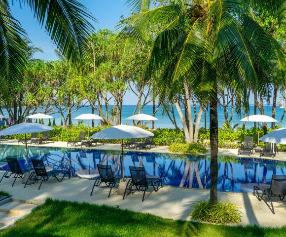 Tropical poolside oasis with palm trees and white umbrellas, overlooking a serene blue ocean. Relaxing lounge chairs line the pool, evoking a tranquil vibe.