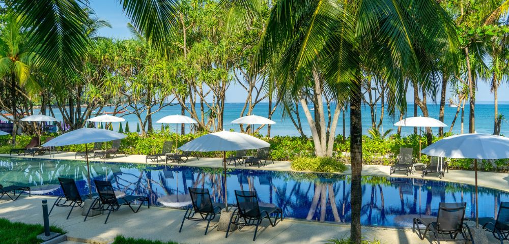 Tropical poolside oasis with palm trees and white umbrellas, overlooking a serene blue ocean. Relaxing lounge chairs line the pool, evoking a tranquil vibe.