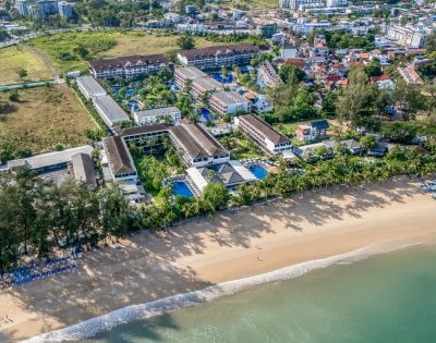 Aerial view of a coastal resort with white sand beach and turquoise sea, surrounded by lush greenery and buildings, evoking a serene and inviting atmosphere.