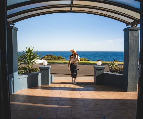 A woman in a sun hat walks towards a building entrance overlooking the ocean. The scene is bright and tranquil, with a clear blue sky and sea.