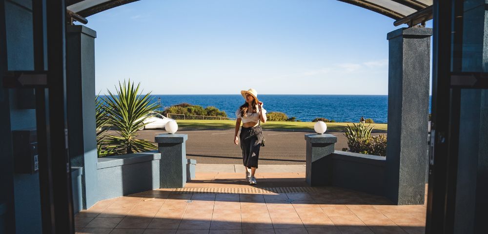 A woman in a sun hat walks towards a building entrance overlooking the ocean. The scene is bright and tranquil, with a clear blue sky and sea.
