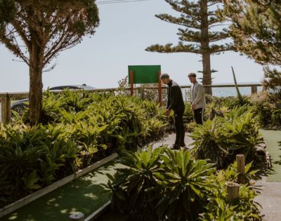 Two people play mini-golf on a sunlit course, surrounded by lush green plants and trees. A blue sky and ocean view create a serene backdrop.