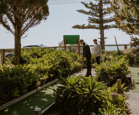 Two people play mini-golf on a sunlit course, surrounded by lush green plants and trees. A blue sky and ocean view create a serene backdrop.