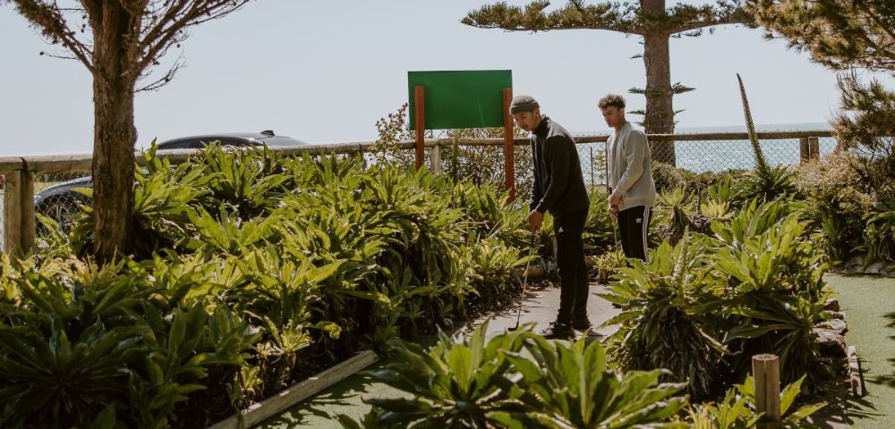 Two people play mini-golf on a sunlit course, surrounded by lush green plants and trees. A blue sky and ocean view create a serene backdrop.