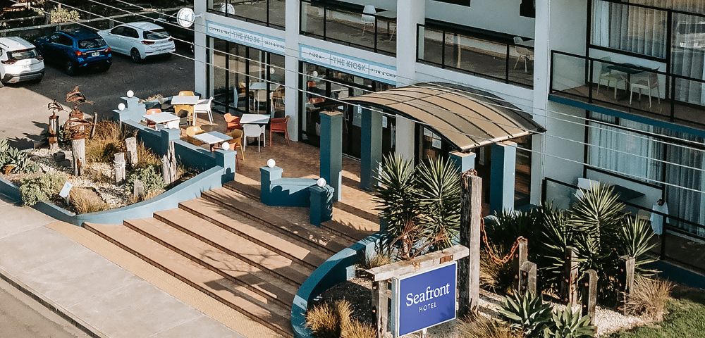 Aerial view of Seafront Hotel entrance with wide, inviting steps and a modern canopy. Surrounded by lush plants and parked cars create a welcoming, relaxed vibe.