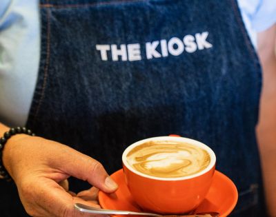 Close-up of a person holding a cappuccino in a red cup. They're wearing a dark apron with 