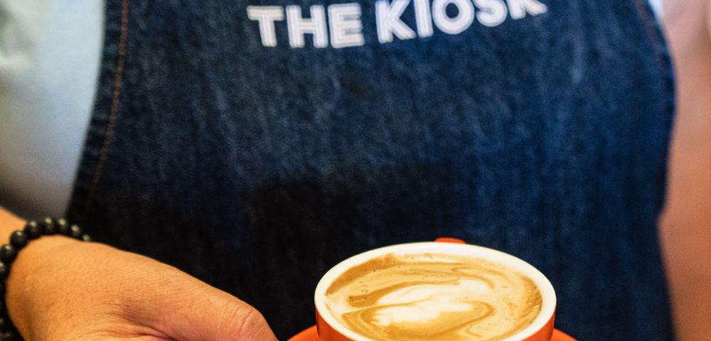 Close-up of a person holding a cappuccino in a red cup. They're wearing a dark apron with 