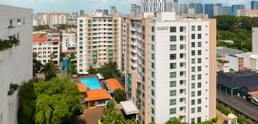 Aerial view of a modern apartment complex with a central orange-roofed pool area surrounded by trees. City skyline and blue sky in the background.
