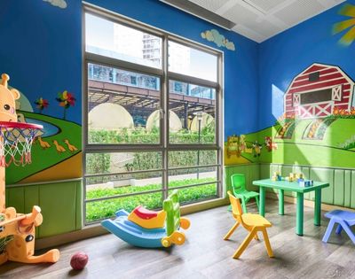 Bright playroom with farm-themed murals, featuring a red barn, green hills, and animals. Colorful toys, chairs, and a small table are scattered around. Large window with outdoor view.