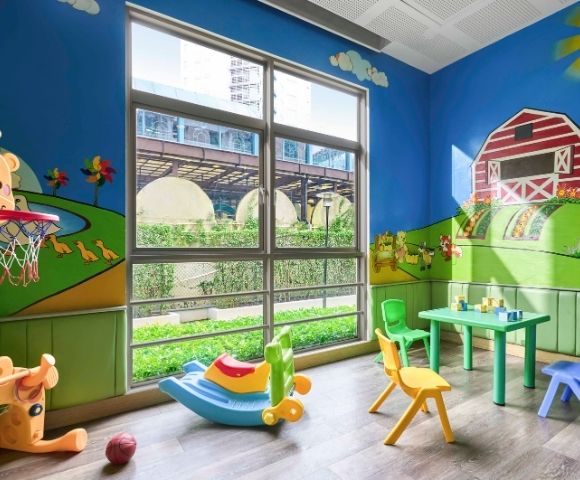 Bright playroom with farm-themed murals, featuring a red barn, green hills, and animals. Colorful toys, chairs, and a small table are scattered around. Large window with outdoor view.