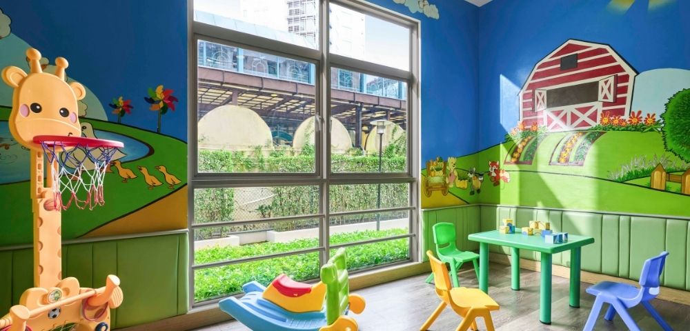 Bright playroom with farm-themed murals, featuring a red barn, green hills, and animals. Colorful toys, chairs, and a small table are scattered around. Large window with outdoor view.