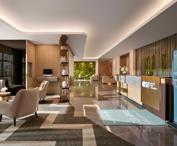 Modern hotel lobby with sleek design, warm lighting, and earth tones. Features plush seating, marble floors, a reception desk, and a green wall backdrop.