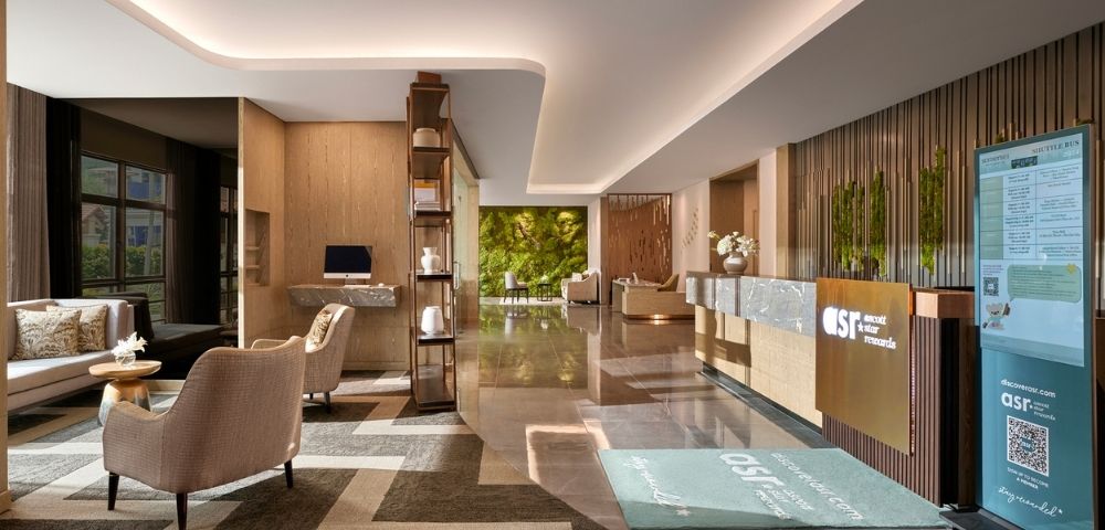 Modern hotel lobby with sleek design, warm lighting, and earth tones. Features plush seating, marble floors, a reception desk, and a green wall backdrop.