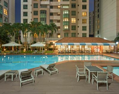 Outdoor pool area in front of a tall apartment building, with lounge chairs and tables on a deck. Palm trees and ambient lighting create a relaxing evening atmosphere.