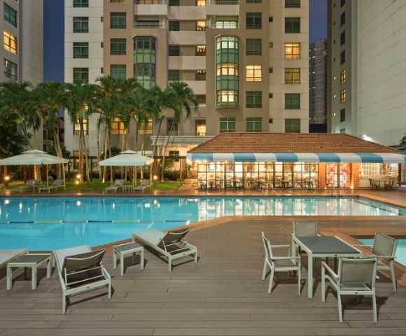 Outdoor pool area in front of a tall apartment building, with lounge chairs and tables on a deck. Palm trees and ambient lighting create a relaxing evening atmosphere.