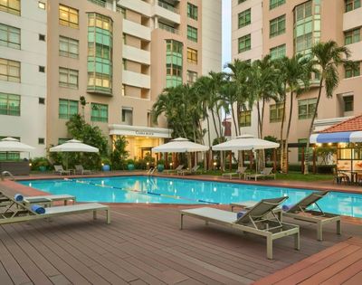 Outdoor pool area surrounded by lounge chairs and umbrellas. Tall buildings and palm trees in the background create a relaxing, resort-like atmosphere.