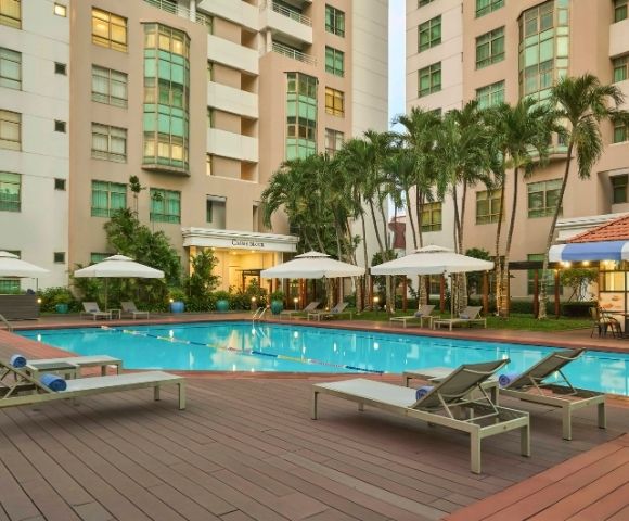 Outdoor pool area surrounded by lounge chairs and umbrellas. Tall buildings and palm trees in the background create a relaxing, resort-like atmosphere.