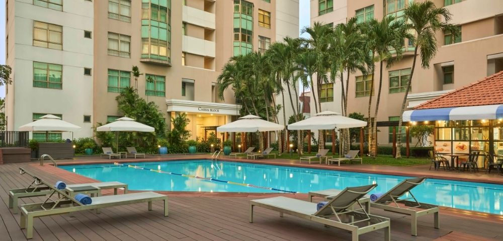 Outdoor pool area surrounded by lounge chairs and umbrellas. Tall buildings and palm trees in the background create a relaxing, resort-like atmosphere.