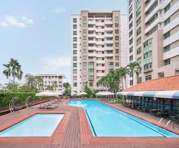 A modern outdoor swimming pool area with a smaller pool, surrounded by high-rise buildings and palm trees. Lounge chairs and umbrellas offer a relaxing vibe.