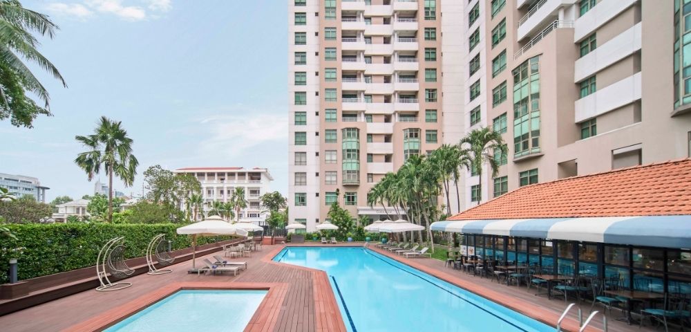 A modern outdoor swimming pool area with a smaller pool, surrounded by high-rise buildings and palm trees. Lounge chairs and umbrellas offer a relaxing vibe.
