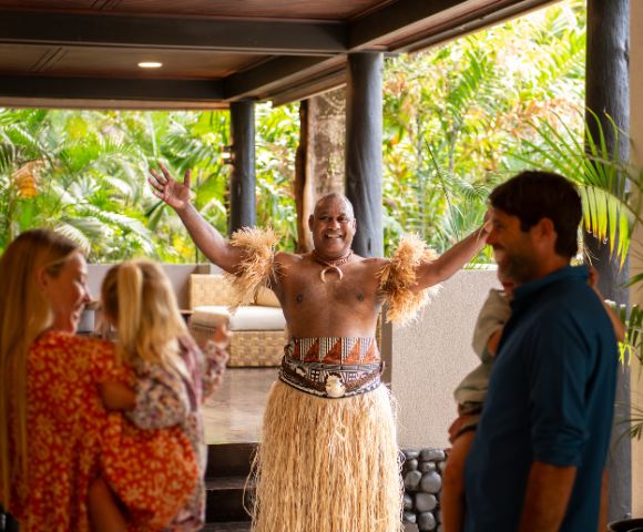 Dancer in traditional attire with grass skirt and arm bands joyfully welcomes a family at a tropical resort entrance, surrounded by lush greenery.