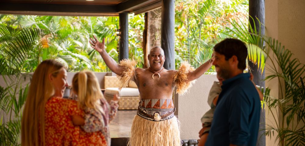 Dancer in traditional attire with grass skirt and arm bands joyfully welcomes a family at a tropical resort entrance, surrounded by lush greenery.