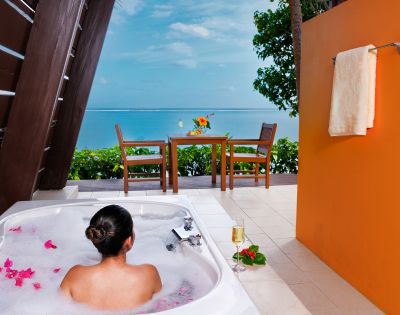 A person relaxes in a bubble-filled outdoor tub with pink petals, overlooking a vibrant sea view. A table with flowers and a towel on an orange wall enhance the tropical ambiance.