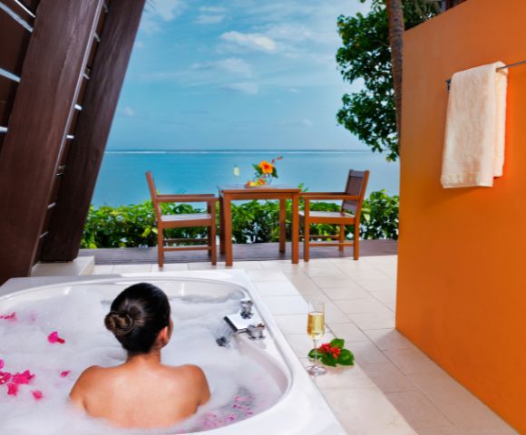A person relaxes in a bubble-filled outdoor tub with pink petals, overlooking a vibrant sea view. A table with flowers and a towel on an orange wall enhance the tropical ambiance.