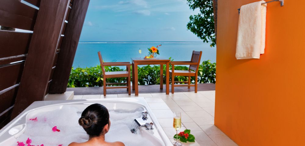 A person relaxes in a bubble-filled outdoor tub with pink petals, overlooking a vibrant sea view. A table with flowers and a towel on an orange wall enhance the tropical ambiance.